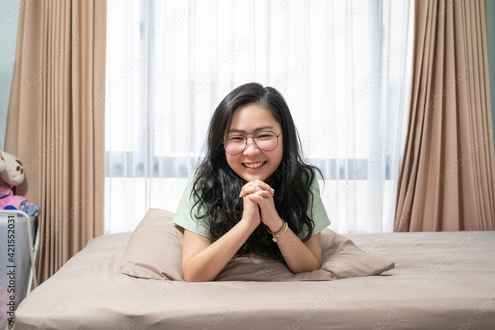 Beautiful Asian woman hold her hands together while lie on her stomach on bed in bedroom with a pastel green - brown color theme. Beautiful Asian woman hold her hands together while lie on her stomach on bed in bedroom with a pastel green - brown color theme.