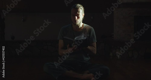 Pensive man reading paper book indoors enlightened by sun rays. Concentrated young yogi meditates alone in dark room looking through breviary slow motion copy space. Mindfulness wellness concept  photo