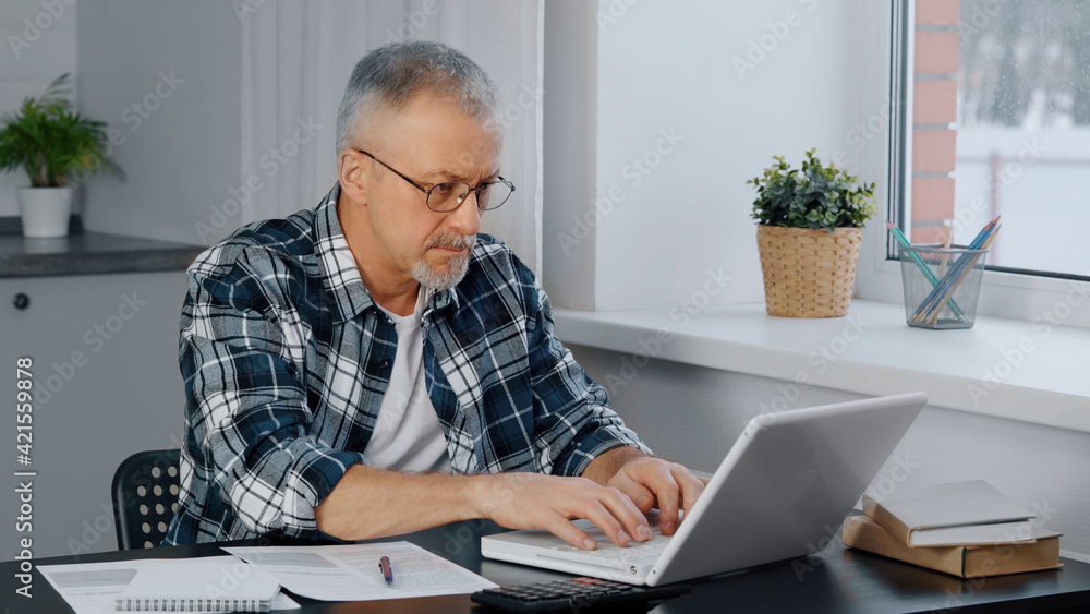 A thoughtful elderly man sits at his laptop, looking through documents. A thoughtful elderly man sits at his laptop, looking through documents.