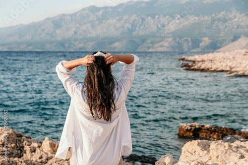 Back view of a Croatian female in a white shirt posing on the rocky coast of the sea