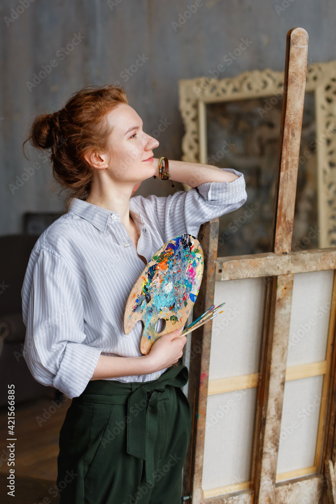 Portrait of a red-haired professional artist with a brush and palette, writing on canvas in the studio. A female artist draws in the workplace. Portrait of a red-haired professional artist with a brush and palette, writing on canvas in the studio. A female artist draws in the workplace.