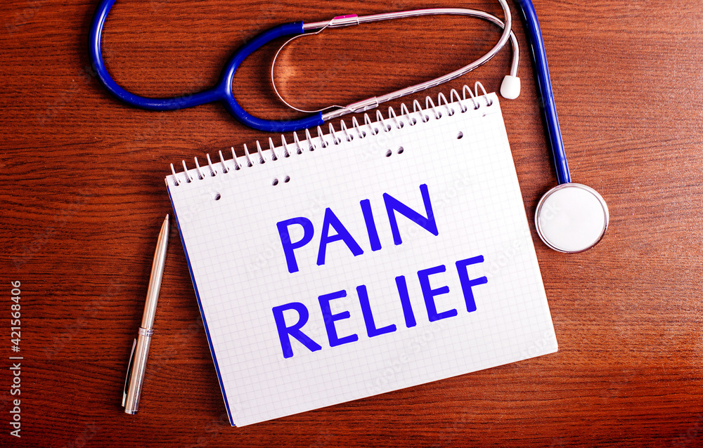 On a wooden table are a pen, a stethoscope, and a notebook labeled PAIN RELIEF. Medical concept On a wooden table are a pen, a stethoscope, and a notebook labeled PAIN RELIEF. Medical concept