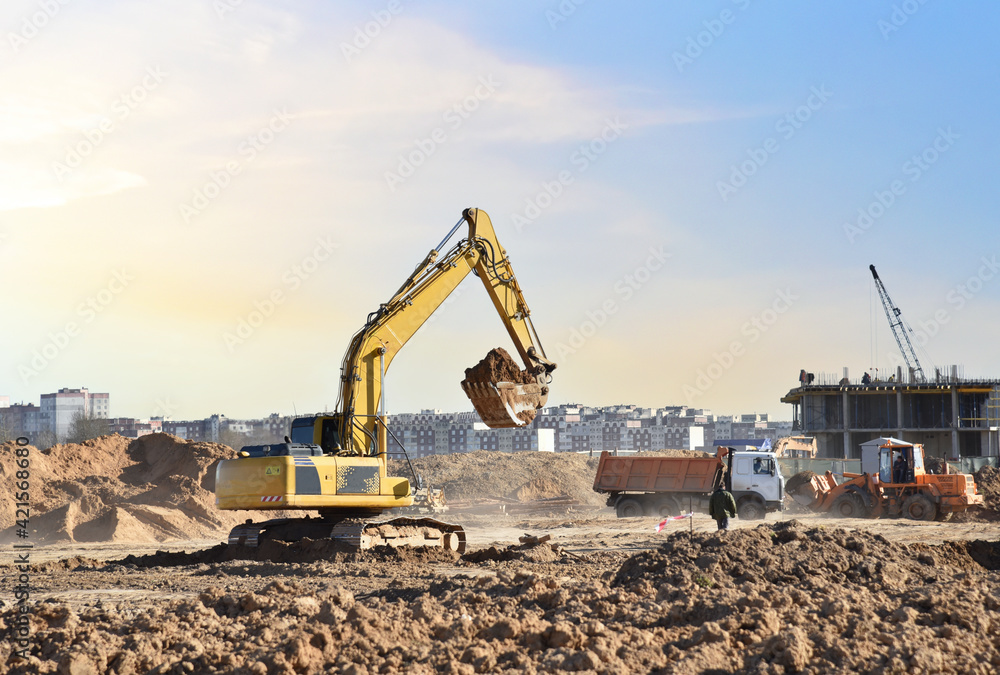 Excavator digging sand and loading into dump truck on construction site. Backhoe digs the ground, foundation for the construction of a new residential building. Tower cranes in action Excavator digging sand and loading into dump truck on construction site. Backhoe digs the ground, foundation for the construction of a new residential building. Tower cranes in action