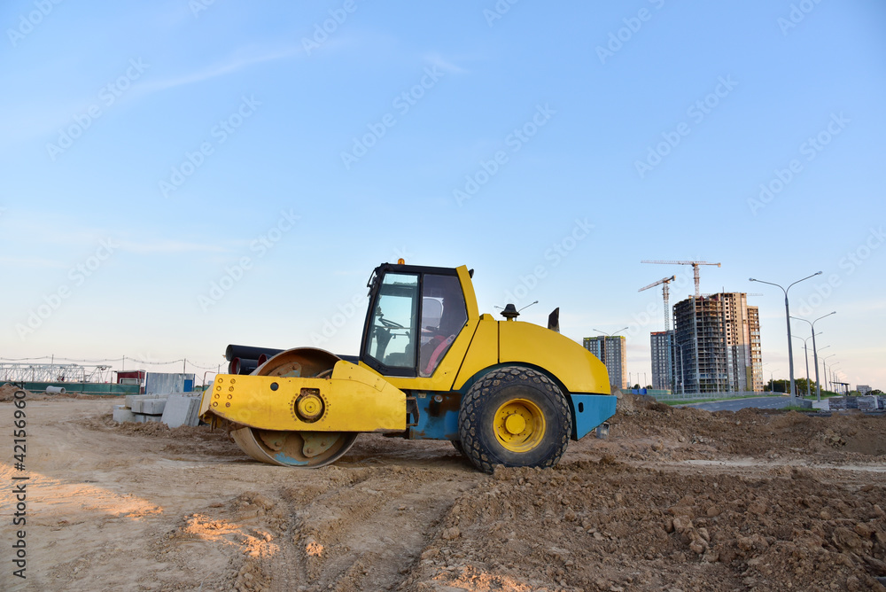Vibro Roller Soil Compactor leveling ground at construction site. Vibration single-cylinder road roller on construction road. Road work for new asphalt laying. Tower cranes build high-rise buildings