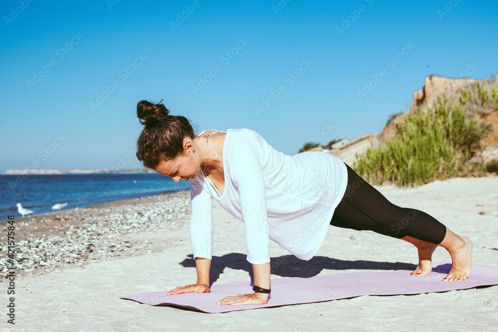 Young sporty woman doing plank or pushups on fitness mat on the beach near ocean in morning, wearing white cloth. Outdoor training, practicing yoga workout in summer. Healthy lifestyle concept. 
