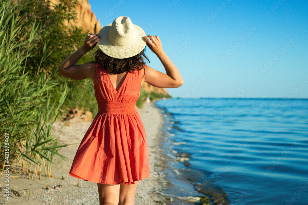 Back view of brunette woman walking along beach in summer straw hat and coral dress on blue ocean and sandy mountains background. Travel, tourism, exploring new routes, enjoying vacation concept