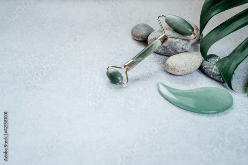 Facial massage tools: jade guasha massage roller and scraper with pebble stones and tropical monstera leaf, SPA relax, face and skin care background