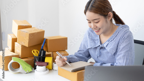 Young Asian woman is writing down the customer's details and addresses on the notebook in order to prepare for shipping according to the information, New kind of business for young, Sell online.