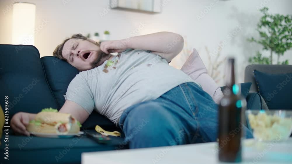 Lazy obese bachelor in dirty clothes sleeping on sofa at home among hot dogs Lazy obese bachelor in dirty clothes sleeping on sofa at home among hot dogs