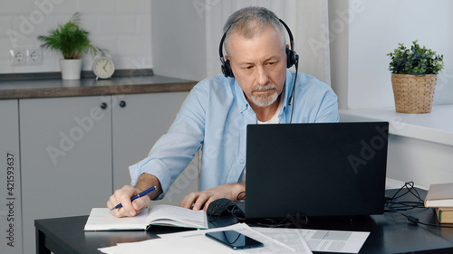 An elderly man wearing headphones sits at a laptop and makes notes.