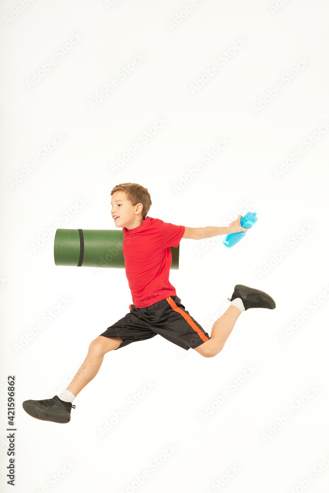 Cute male kid with yoga mat and bottle of water jumping in studio Cute male kid with yoga mat and bottle of water jumping in studio