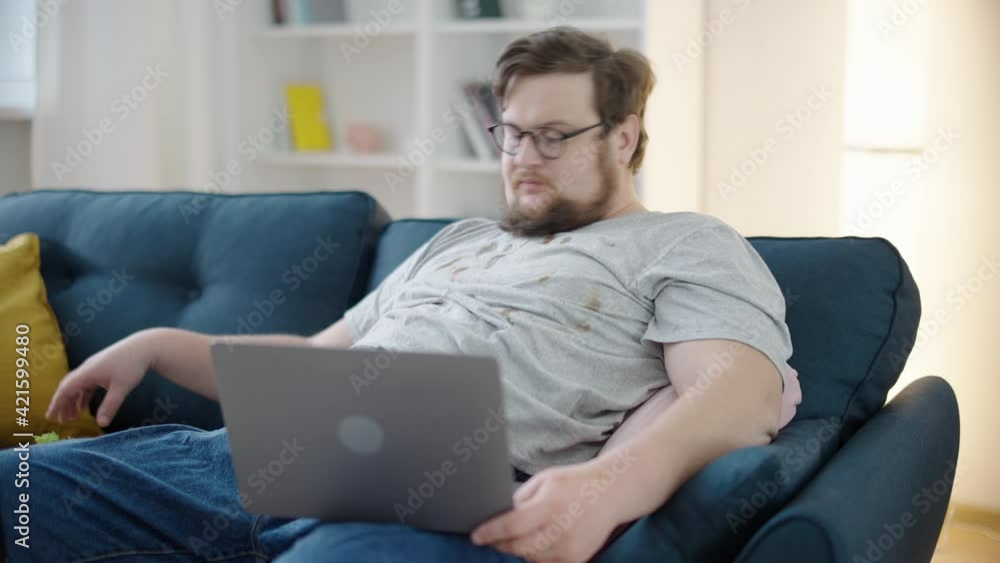 Messy oversize man in dirty T-shirt eating sandwich, watching video on laptop Messy oversize man in dirty T-shirt eating sandwich, watching video on laptop