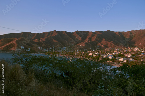 Hermoso atardecer en la ciudad de Taganga, un pueblo de pescadores con muchos botes en la orilla de la playa y edificios entre los árboles con un cielo azul claro sin nubes.