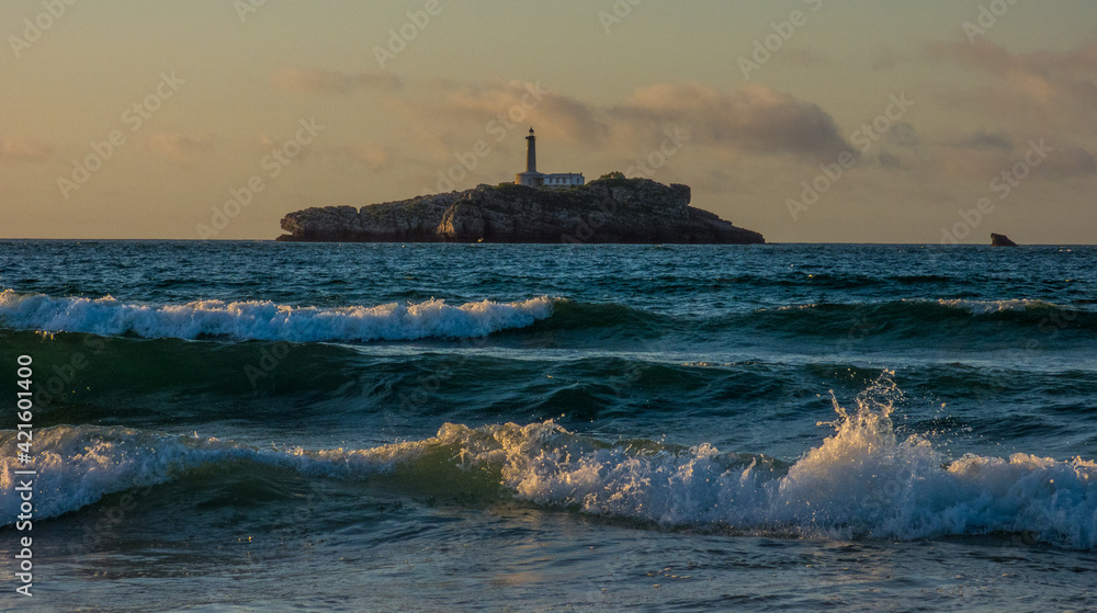 Isla con grandes rocas formando acantilados coronada por un faro y bajo ella las olas de la playa desde la que se hizo la foto