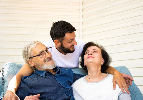 Happy senior couple sitting on sofa while his thankful adult brunette bearded son hugging them with love and care, enjoying weekend family time at home together. Generation and people concept