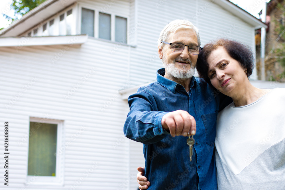 Fototapeta premium Smiling senior couple holding keys to new own housing, mature family bonding to each other and looking at camera, standing in front of big white house.Happy and prosperous old age concept. Copy space