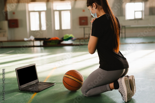Basketball coach wearing medical mask is praying for end of pandemic, in empty sport gym. Online communication, training in front of empty blank laptop computer screen