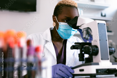Black female scientist in mask in laboratory looking through microscope photo