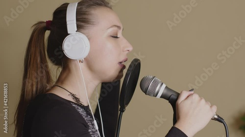 Young beautiful female musicant in headphones singing in professional microphone. Pretty woman singing with sensuous emotions. Vocal online lessons, music vlog, live streaming, broadcasting concept