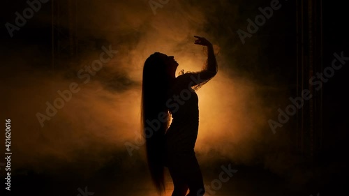 Side view silhouette of young woman dancing in dark smoky studio against background of yellow lights. Female with long hair performs sensual movements with hands touching body. Close up. Slow motion. photo