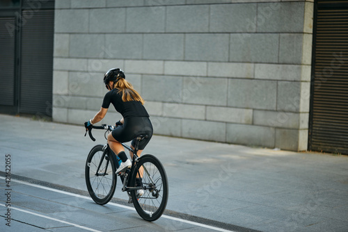 Rear view of professional female cyclist in black cycling garment and protective gear riding bicycle in city, passing buildings while training outdoors on a daytime