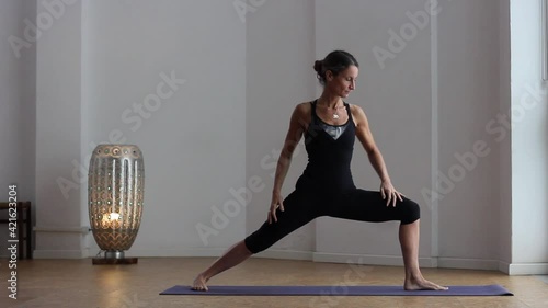 Female yogi doing warrior pose alone in studio with lamp by white background. Middle aged woman practicing virabhadrasana two indoors photo