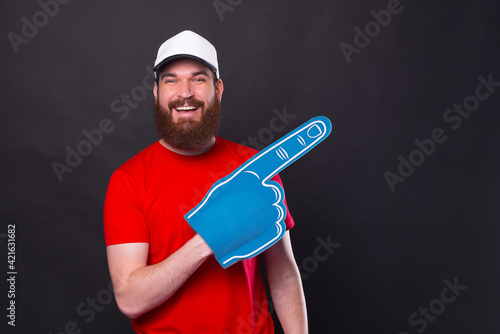 Cheerful portrait of young bearded man in red t-shirt pointing away with fan foam glove photo