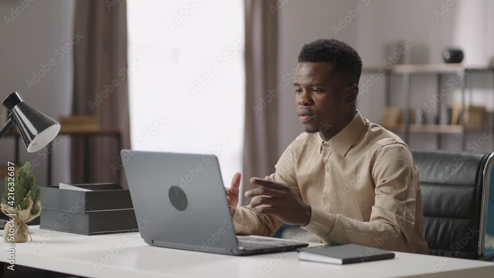 young black man is communicating by video chat in laptop, working from his home or office, medium portrait indoor