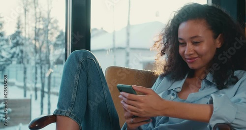 Beautiful happy young African American student woman with curly hair using smart phone in comfortable cozy chair at home photo