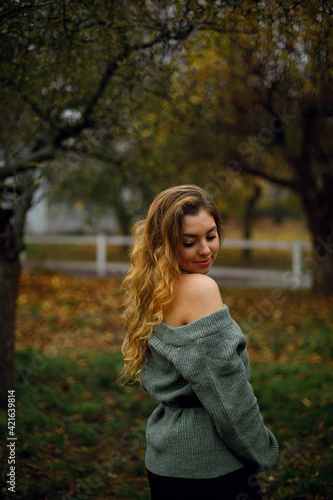 portrait of young beautiful Caucasian woman with wavy hair. a girl in a warm sweater stands with an open shoulder in the middle of the garden or on her ranch. 