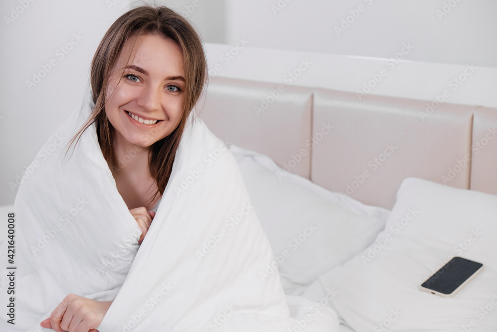 Pretty young smiling Caucasian woman waking up in the morning and stretching in her bed and looking content. Covered in white linen. Pretty young smiling Caucasian woman waking up in the morning and stretching in her bed and looking content. Covered in white linen.