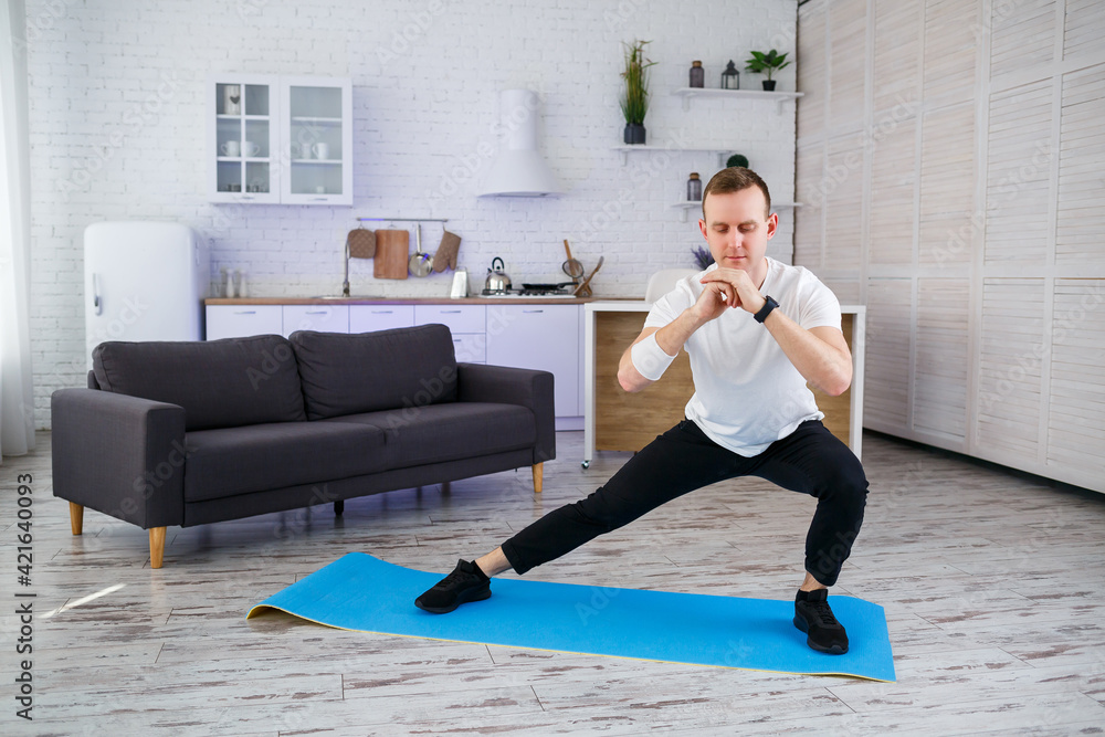 Obraz premium A strong athletic man in a T-shirt does exercises in his spacious apartment with a minimalistic interior. Healthy lifestyle during the quarantine period. Fitness at home