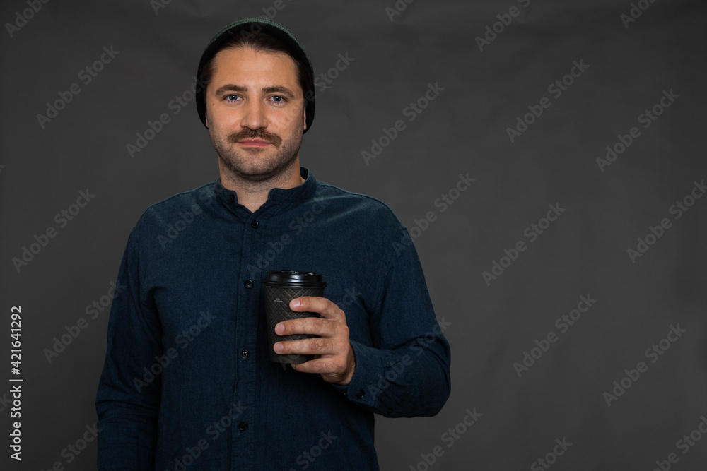 Handsome unshaven guy in knitted cap posing with disposable cup looking at camera on gray studio backdrop copy space. Morning wake-up coffee, take-away coffee, coffee shop or snack bar advertising Handsome unshaven guy in knitted cap posing with disposable cup looking at camera on gray studio backdrop copy space. Morning wake-up coffee, take-away coffee, coffee shop or snack bar advertising
