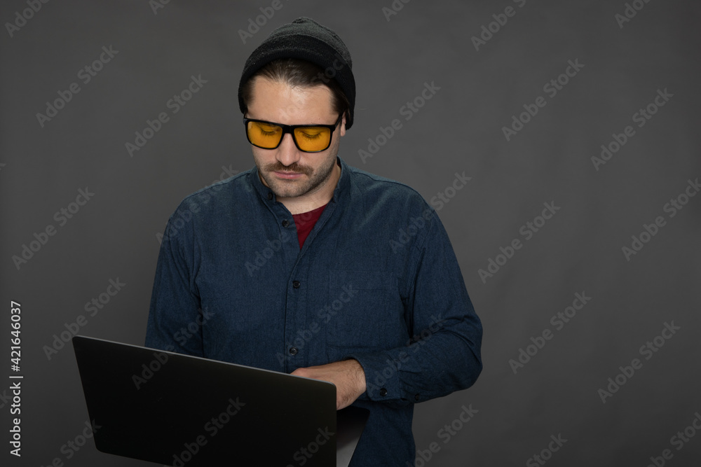 Handsome unshaven guy in knitted cap and yellow glasses standing up and working with laptop on gray studio backdrop copy space.