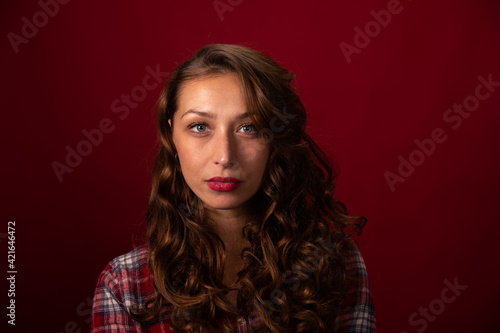 Close up portrait of Beautiful curly long hairy model in plaid shirt posing on red studio backdrop looking at camera. Pretty girl, women's beauty standards, cosmetics advertising copy space