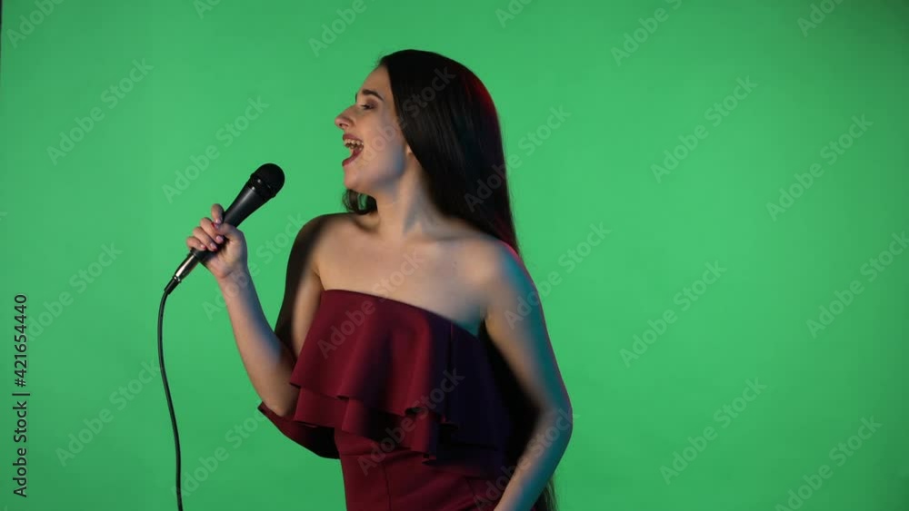 Portrait of beautiful young woman illuminated by blue neon light sings song into microphone. Model with long hair in red dress posing on green screen in studio. Side view. Slow motion. Close up. Portrait of beautiful young woman illuminated by blue neon light sings song into microphone. Model with long hair in red dress posing on green screen in studio. Side view. Slow motion. Close up.