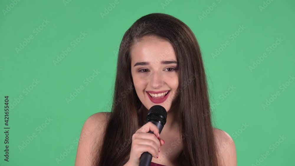 Portrait of beautiful young woman coquettishly looking at camera and sings song into microphone. Model with long hair in red dress posing on green screen in the studio. Slow motion. Close up. Portrait of beautiful young woman coquettishly looking at camera and sings song into microphone. Model with long hair in red dress posing on green screen in the studio. Slow motion. Close up.