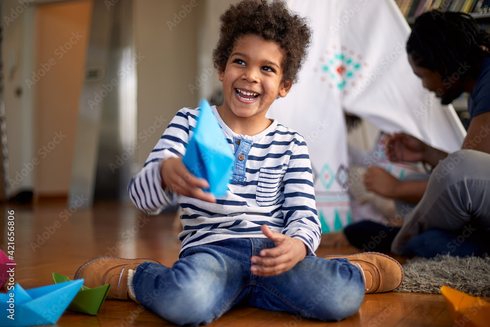 A cute little boy playing with paper boats on the floor and posing for a photo while spending a time with his family at home. Family, home, playtime A cute little boy playing with paper boats on the floor and posing for a photo while spending a time with his family at home. Family, home, playtime
