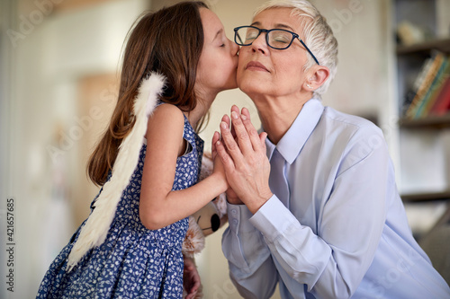 A little girl with angel wings is kissing and exchanging emotions with her grandmother at home. Family, home, love, playtime