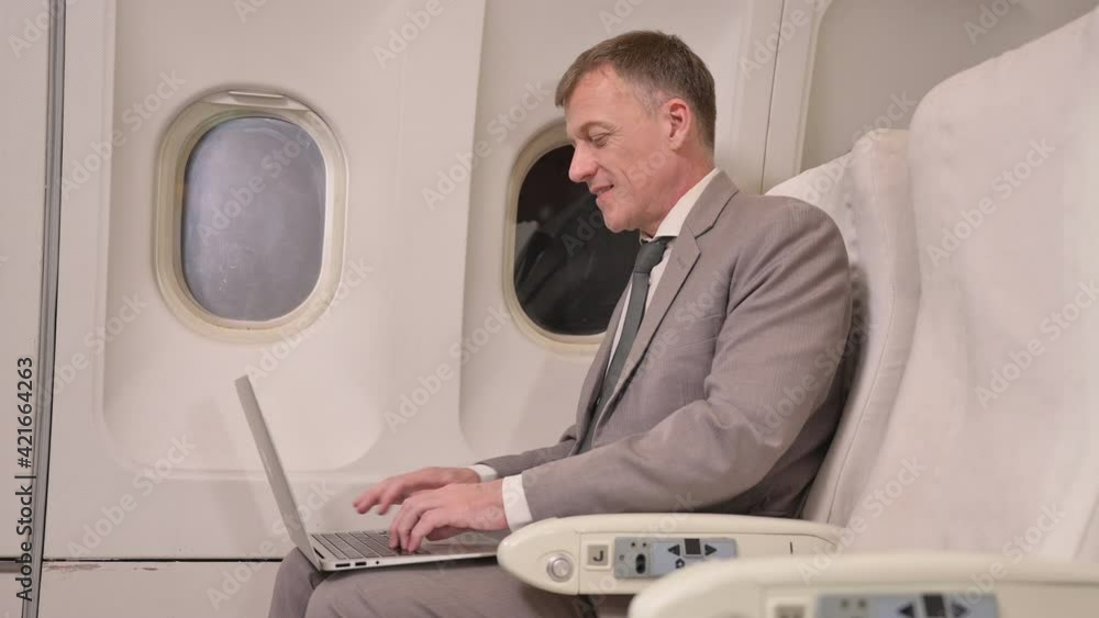 Senior professional businessman using computer laptop on airplane. Happy handsome business man in suit sitting and smiling near window with typing on keyboard while using laptop on first class flight Senior professional businessman using computer laptop on airplane. Happy handsome business man in suit sitting and smiling near window with typing on keyboard while using laptop on first class flight