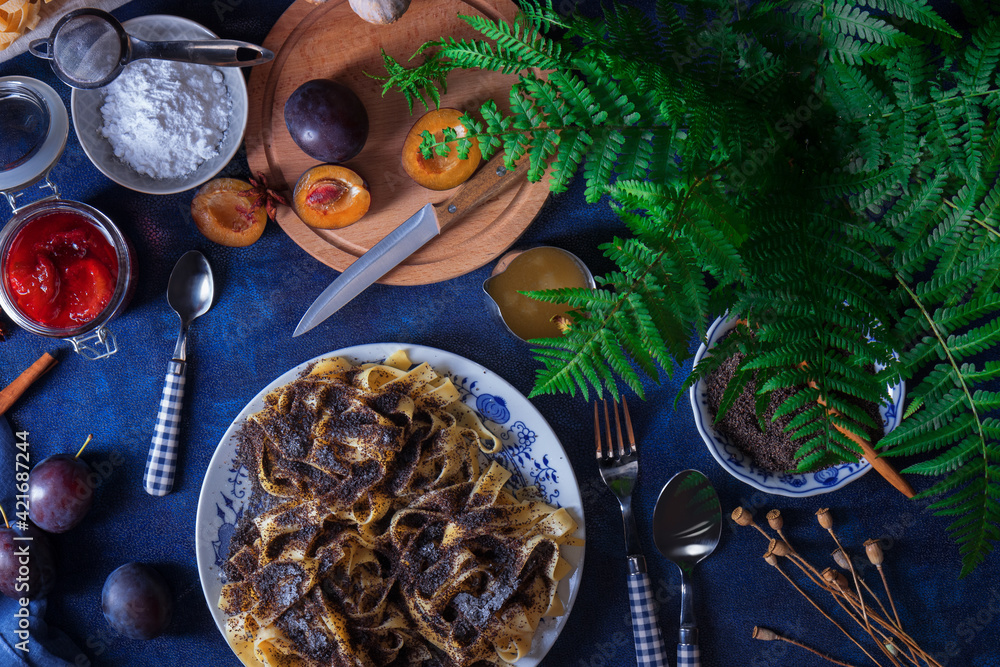 Sweet pasta dessert, noodles with poppy seeds, plum compote from fresh plums, dark background, blue table decorated with fresh flowers. Dry whole poppy plant in background.