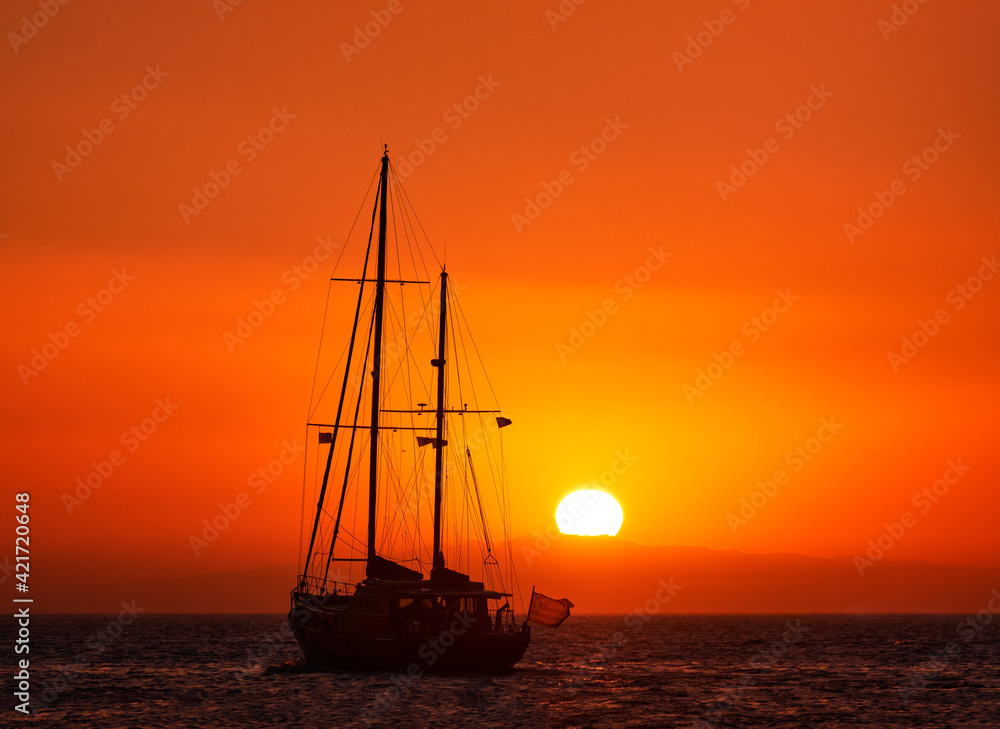 Silhouette of sailing boat with sails down against sun at sunset, sun glare on sea waters. Romantic seascape. Sailboat go to sea