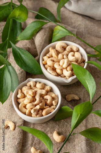 Cashew nuts in two white cups. Fabric background. Nearby cashews and green leaves. View from above.