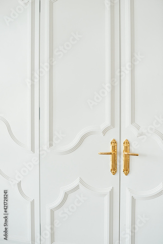 Close-up of white baroque doors with classic golden handles and ornaments. Beauty in details..Beautiful entrance to an expensive apartment. Copy space.