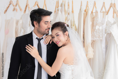 Mix race wedding concept, Happy couple spent time together while choosing or fitting wedding dress and tuxedo in fitting room, beautiful Asian woman hold or hug her man with passion and love