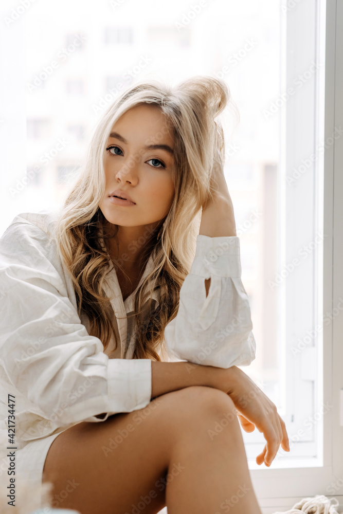 Young blonde woman with long wavy hair, wearing white shirt, sitting in soft window light. Young blonde woman with long wavy hair, wearing white shirt, sitting in soft window light.