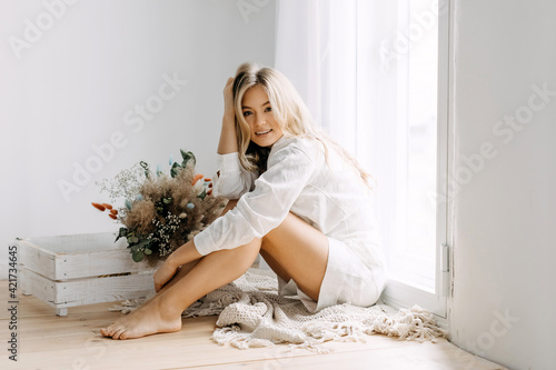 Young blonde woman with long wavy hair, indoors, sitting on the floor by the window.