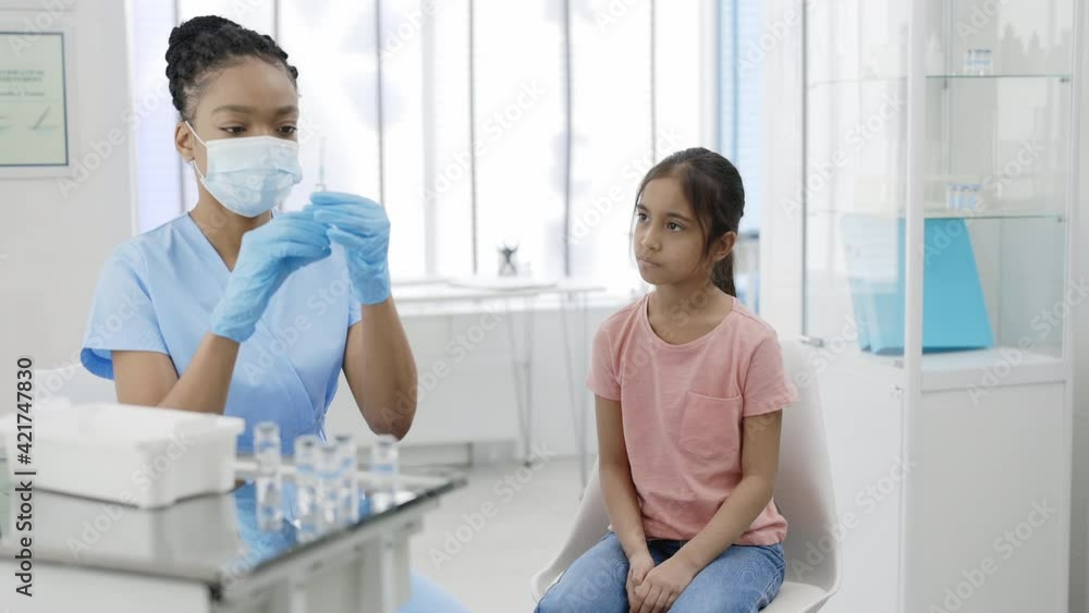 Crop view of nurse in protective medical mask and safety gloves prepering syringe with vaccine for young girl. Kid waiting for vaccination while sitting on chair in hospital. Crop view of nurse in protective medical mask and safety gloves prepering syringe with vaccine for young girl. Kid waiting for vaccination while sitting on chair in hospital.