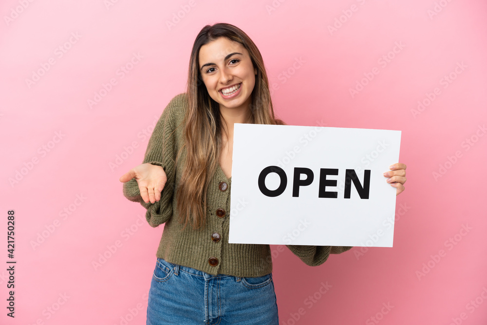 Young caucasian woman isolated on pink background holding a placard with text OPEN and pointing it Young caucasian woman isolated on pink background holding a placard with text OPEN and pointing it