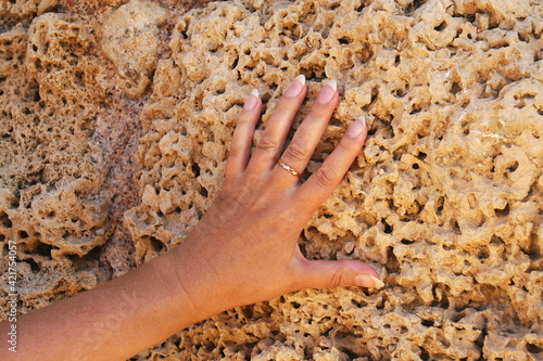 A tanned female left hand with a forearm with a gold ring on the middle finger against the background of an old weathered sand-colored stone.